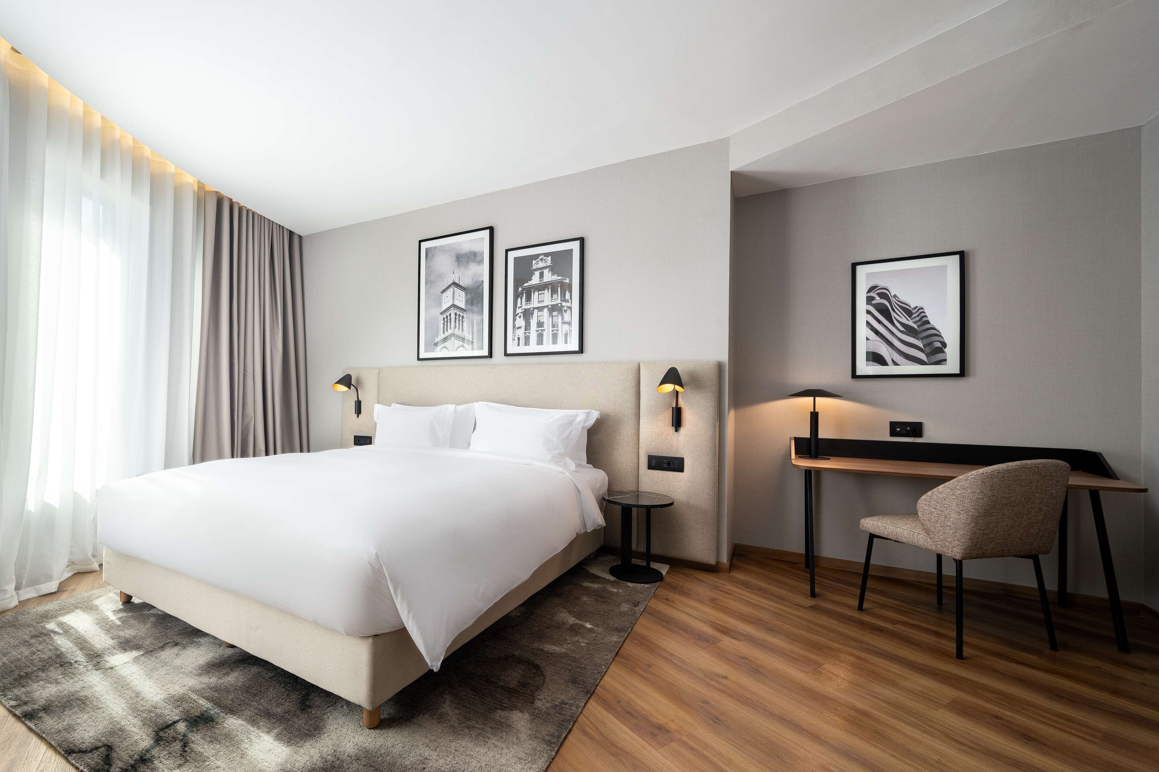 A hotel room with a large bed, a desk with a chair, and two wall-mounted lamps.