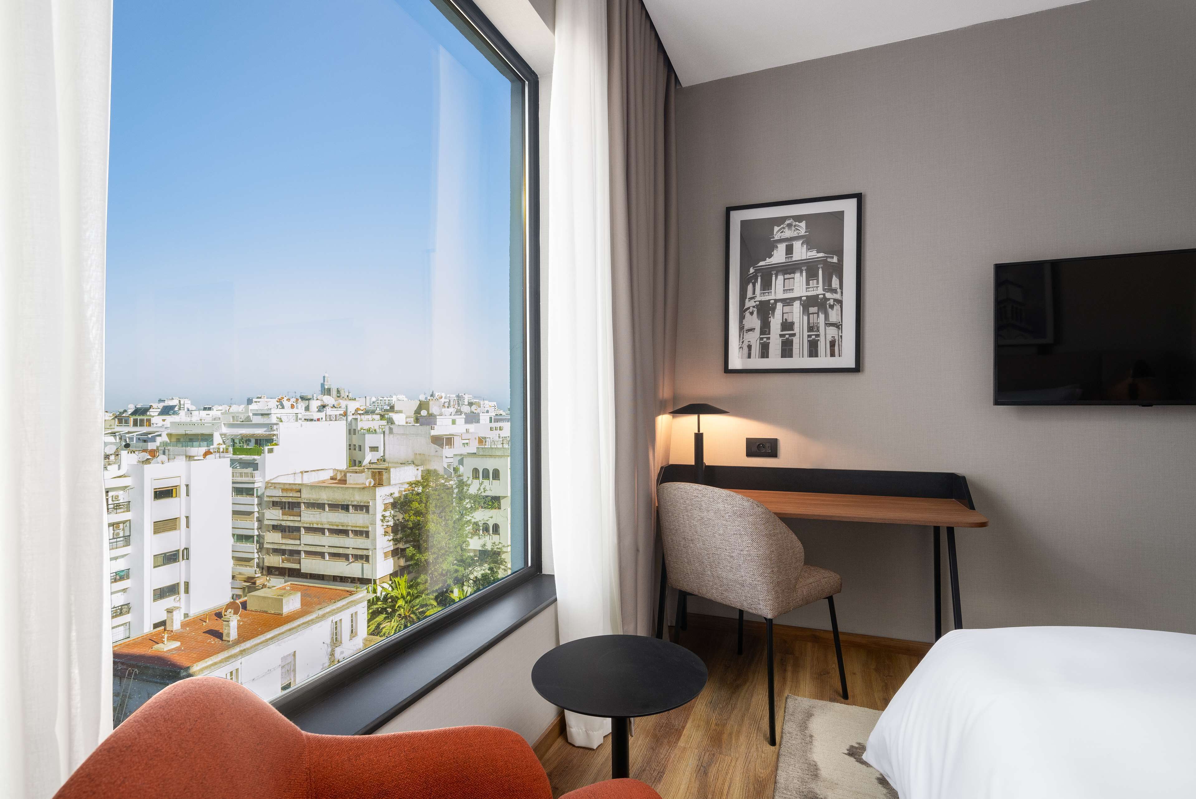 A hotel room with a large window, a desk, a chair, and a bed.