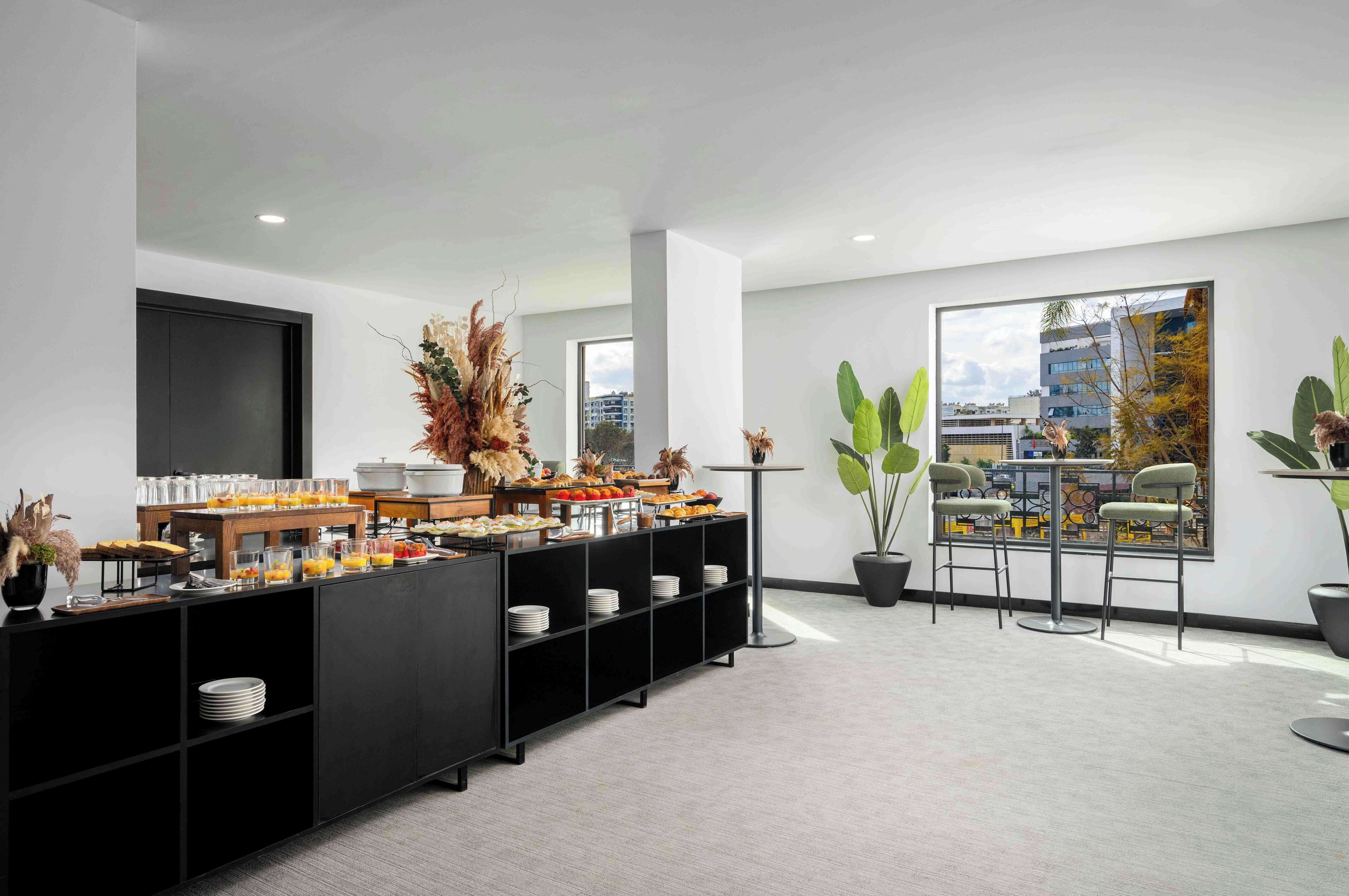 A modern hotel room with a large window, a black buffet table with various food items, a potted plant, and a painting on the wall.