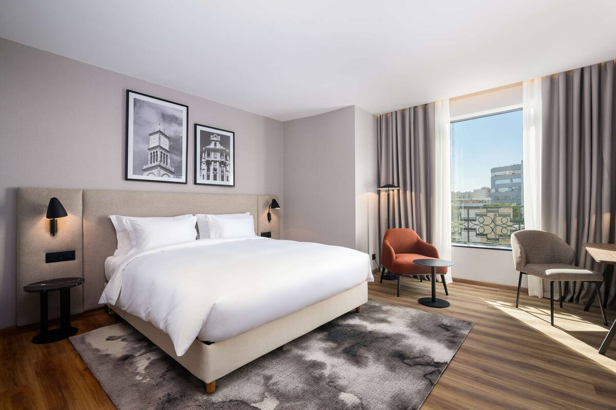Une chambre d’hôtel moderne, dotée d’un grand lit, de deux tables de chevet, d’une chaise, d’une petite table et offrant une vue sur la ville grâce à de grandes fenêtres.