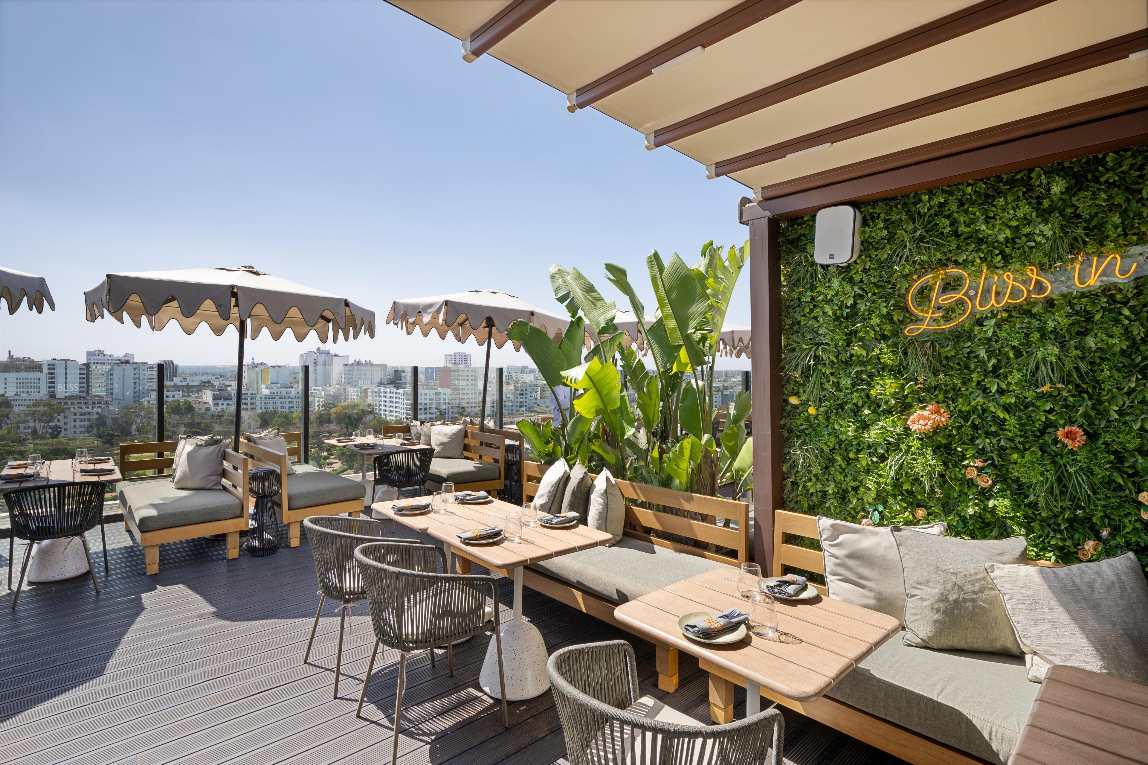 An outdoor seating area with tables and chairs, a view of the city, and a wall with greenery and a neon sign.