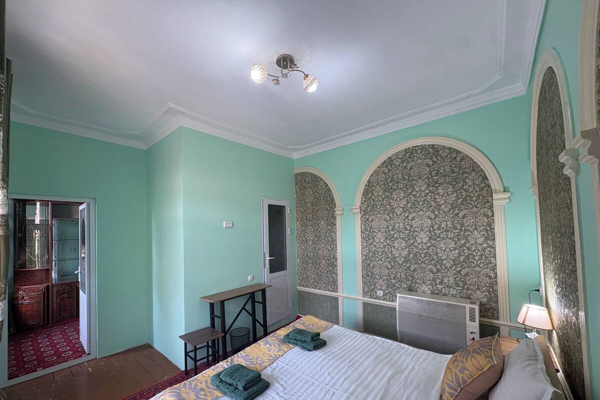 A bedroom with a bed, a desk, and a mirror. There is a door leading to another room.