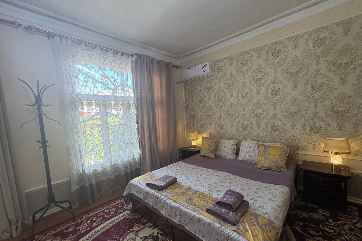 A hotel room with a large bed, two bedside lamps, a window with curtains, a coat rack, and a floral-patterned carpet.