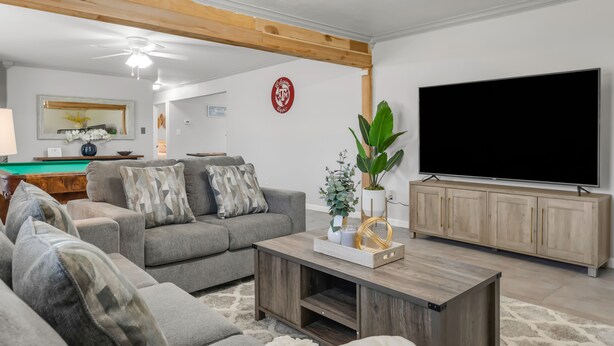 A living room with a grey sofa, a wooden coffee table, a flat-screen TV, and a pool table in the background. - A&M SPORTS DAY, POOL TABLE, AND BBQ (College Station)