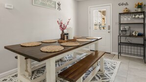 A dining area with a wooden table, woven placemats, and benches. There is a coffee station with a shelf holding mugs and a coffee maker. - A&M SPORTS DAY, POOL TABLE, AND BBQ (College Station)