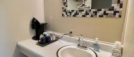 Un bagno con un lavandino bianco, uno specchio con cornice in mosaico bianco e nero, un asciugacapelli e una tazza di caffè sul ripiano.