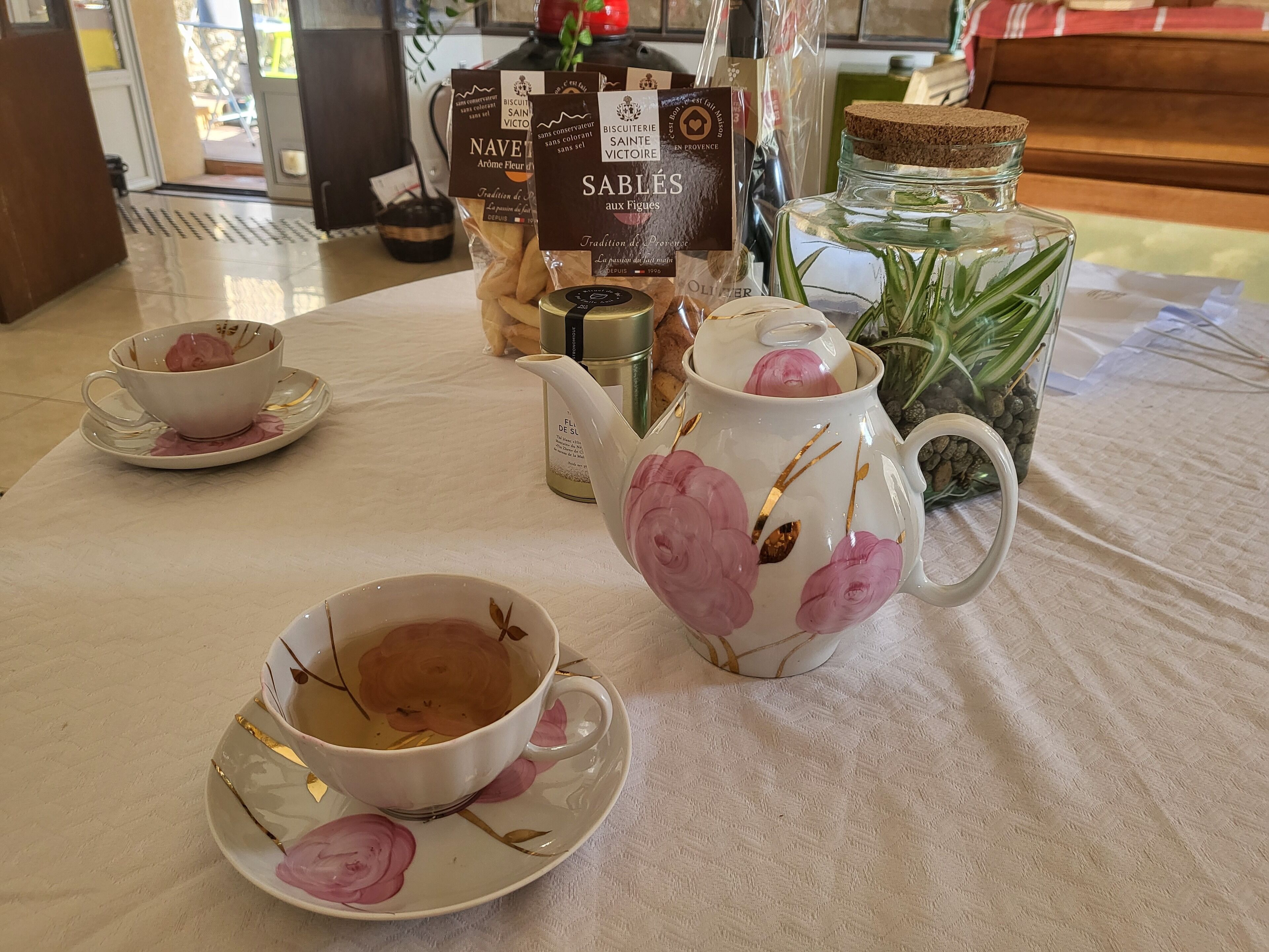Une table dressée avec des tasses à thé et une théière, un bocal de biscuits et une bouteille d’huile d’olive.
