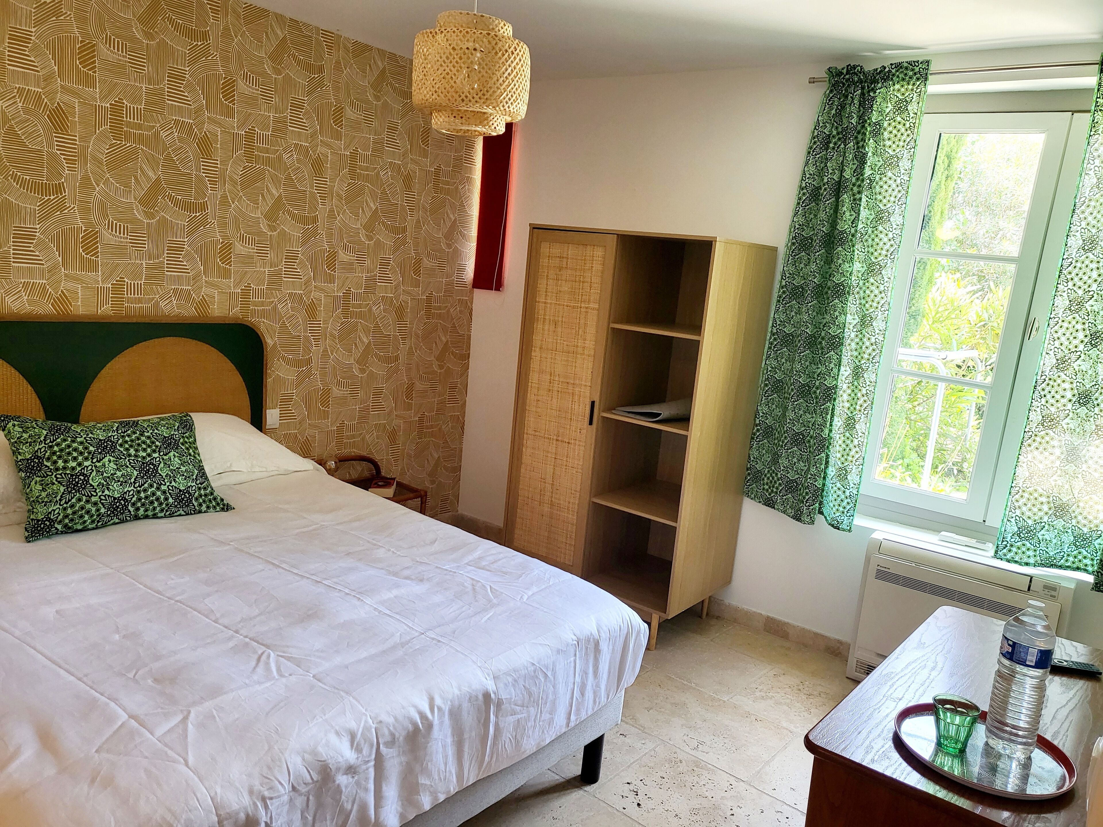 Une chambre à coucher comprenant un lit, une armoire, une fenêtre avec des rideaux et une table de chevet sur laquelle se trouvent une bouteille et un verre.