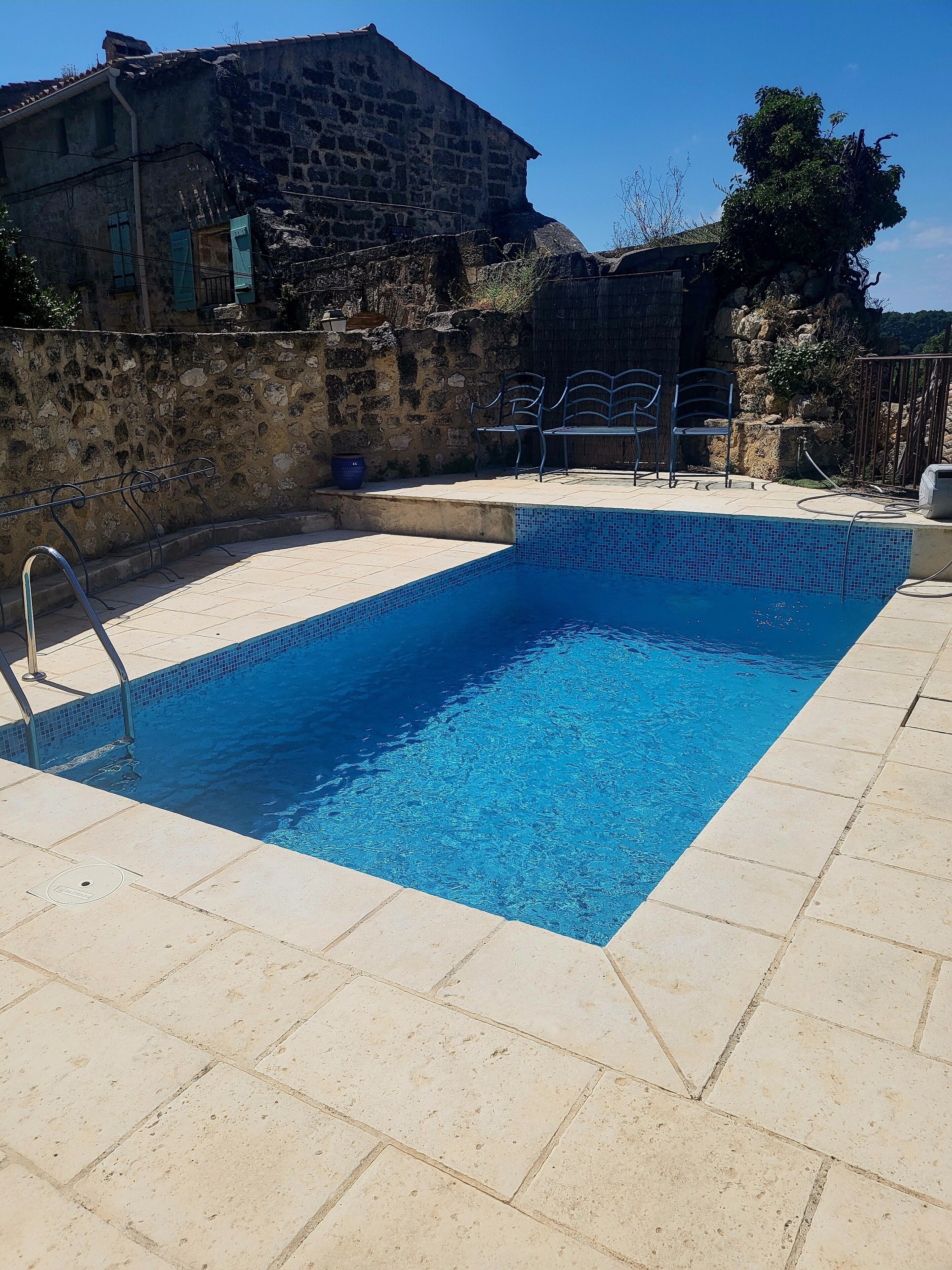 A rectangular swimming pool with a tiled edge, surrounded by a stone wall and a building with a stone facade.