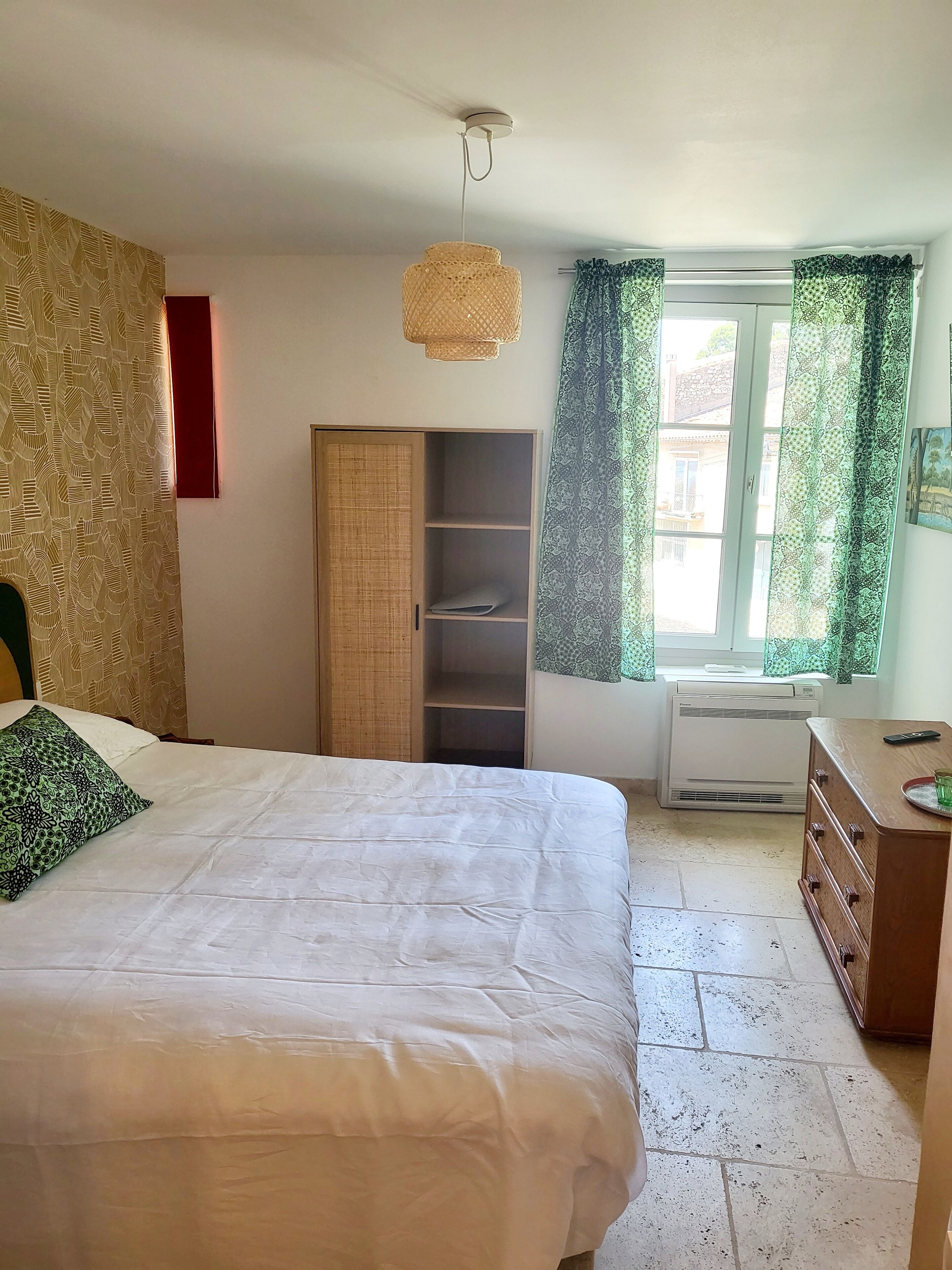 A bedroom with a bed, wardrobe, air conditioning unit, and a window with curtains.