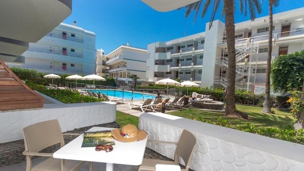 Zona de piscina con sillones de descanso y sombrillas, una mesa con un sombrero y gafas de sol, y vistas a un edificio con varios balcones.