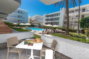 Un espace aménagé au bord de la piscine, avec des chaises longues et des parasols, une table sur laquelle sont posés un chapeau et des lunettes de soleil, et une vue sur un immeuble doté de nombreux balcons.