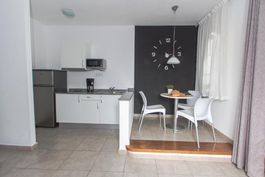 A compact kitchen with white cabinets, a microwave, and a coffee maker. There is a dining area with two white chairs and a small table. A black clock is mounted on the wall.