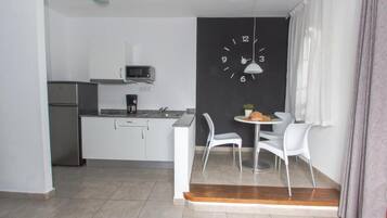 A compact kitchen with white cabinets, a microwave, and a coffee maker. There is a dining area with two white chairs and a small table. A black clock is mounted on the wall.