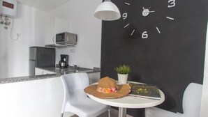 Una cocina moderna con microondas, cafetera y un reloj en la pared. Hay dos sillas blancas y una mesa con un sombrero y una planta.