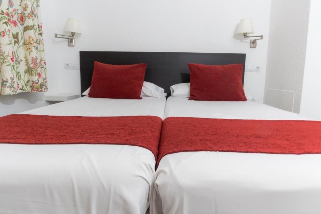 A double bed with white bedding and red accents, two bedside lamps, and a floral curtain.