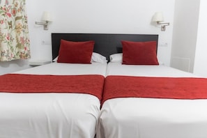 Cama doble con ropa blanca y detalles rojos, dos lámparas de noche y una cortina con motivos florales.