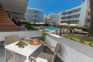 Zona junto a la piscina con sillas de descanso y sombrillas, una mesa blanca con un sombrero y gafas de sol, y una escalera que lleva a un edificio con balcones.