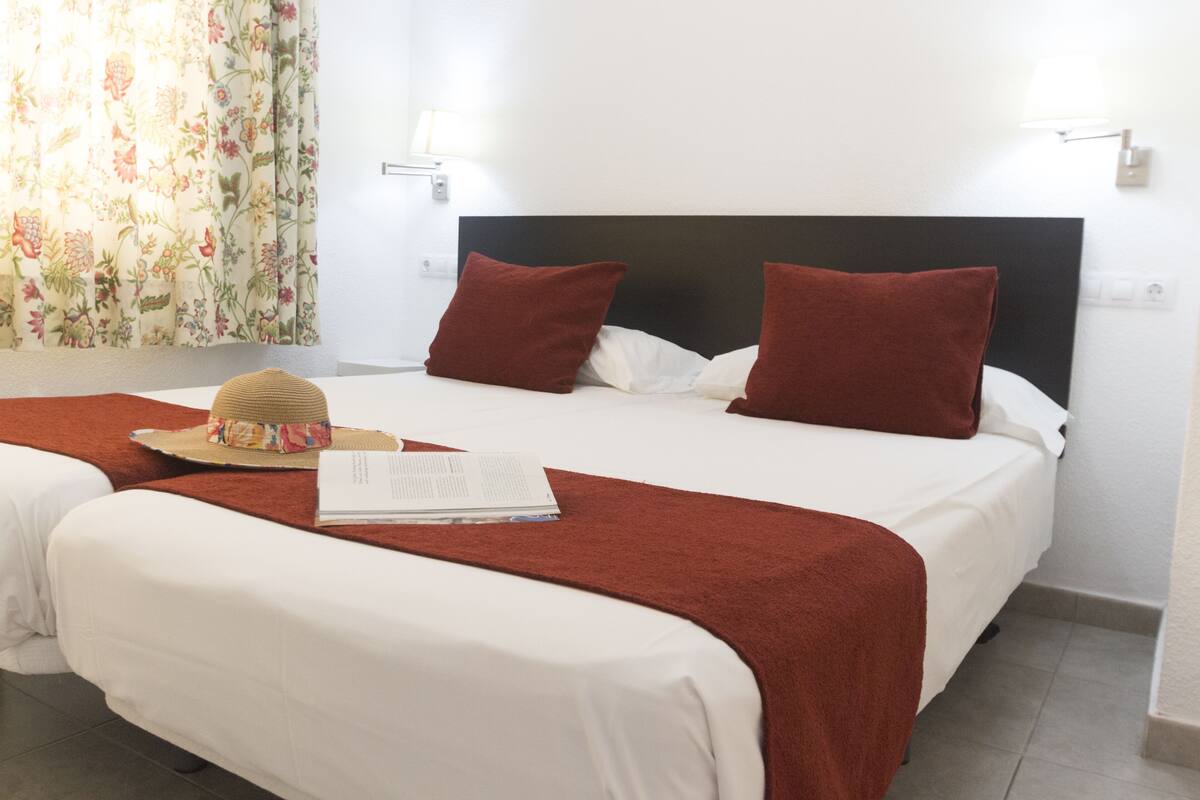 Ein Hotelzimmer mit zwei Betten, beide mit weißer Bettwäsche und roten Kissen. Auf dem Bett links liegen ein Hut und ein Buch.