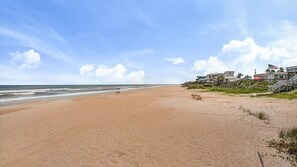 Ein Strand mit blauem Himmel, sandigem Ufer und Gebäuden im Hintergrund.