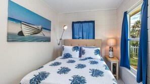 Une chambre à coucher avec un lit, un tableau représentant un bateau, une lampe et une fenêtre avec des rideaux.