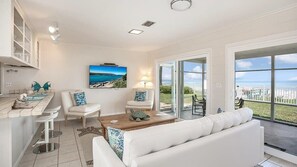 A modern living room with a white sofa, two chairs, a coffee table, and a TV. There is a large window with a view of the ocean and a balcony.