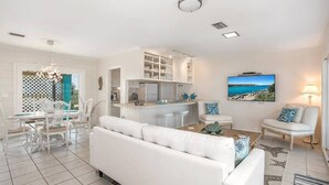 Un salon moderne avec un canapé blanc, un coin repas avec des chaises blanches, une cuisine avec des meubles blancs et un téléviseur diffusant un paysage marin.