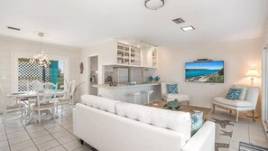 A modern living room with a white sofa, dining area with white chairs, a kitchen with white cabinets, and a TV displaying a coastal scene.