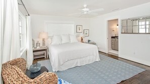 Ein Schlafzimmer mit Bett, Nachttischen, einer Lampe, einem Sessel, einem Fenster mit Jalousien, einem Deckenventilator und einem durch eine offene Tür einsehbaren Badezimmer.