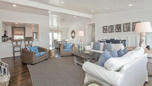 A spacious living area with a white sofa, wicker chairs, and a coffee table. A kitchen is visible in the background with white cabinets and a stone backsplash. - Sandy Chic Home with Oceanfront Deck & Fireplace (Ponte Vedra Beach)