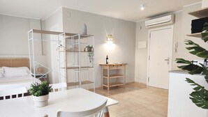 A modern, well-lit room with a bed, white furniture, a wooden side table, and a potted plant. - Mazi Apartments Havana (Mataró)