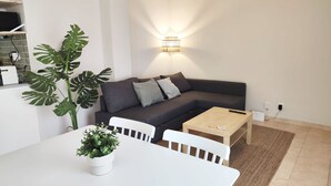 A modern living room with a dark grey sofa, white chairs, a wooden coffee table, and a large green plant. - Mazi Apartments Havana (Mataró)