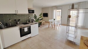 A modern kitchen with white cabinets, a microwave, and a sink. There is a dining area with a table and chairs, and a large plant in the center. - Mazi Apartments Havana (Mataró)