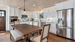 Una cocina moderna con armarios blancos, electrodomésticos de acero inoxidable, una mesa de comedor de madera y una lámpara colgante.