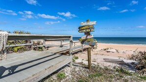 Un pasarelas de madera que conduce a una playa, con un cartel que indica varios destinos.