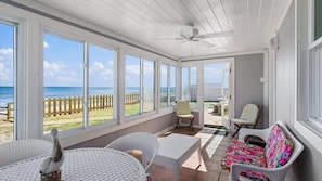 Eine überdachte Terrasse mit großen Fenstern zum Meer, Korbmöbel und einem Deckenventilator.