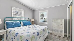 A bedroom with a bed, two bedside tables, a dresser, and two windows with blinds.