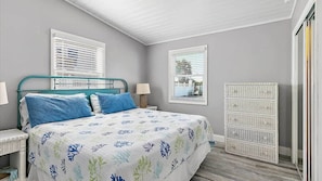 A bedroom with a bed, two bedside tables, a dresser, and two windows with blinds.
