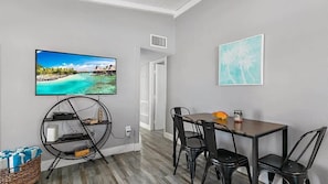 Een kamer met een televisie waarop een tropisch strand te zien is, een eettafel met stoelen en een wandplank met boeken en een lamp.