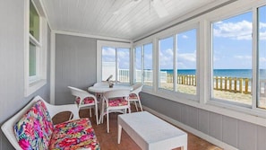 Eine überdachte Terrasse mit Tisch und Stühlen, einer Bank und großen Fenstern mit Blick auf den Strand.