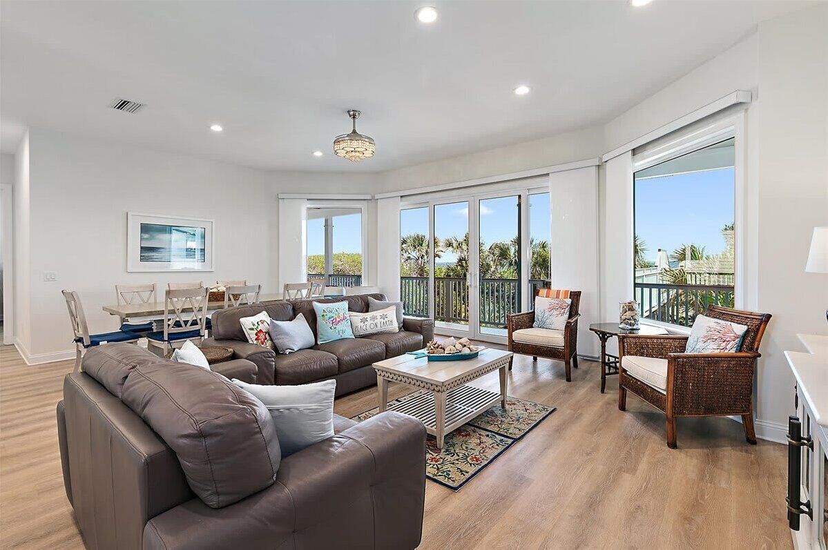 A spacious living room with a brown leather sofa, a coffee table, wicker chairs, and a dining area with a white table and chairs.