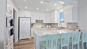 A modern kitchen with white cabinetry, a central island, and a dining area with light blue chairs. - Sandy Toes Home with Hot Tub & Dune Deck (Ponte Vedra Beach)