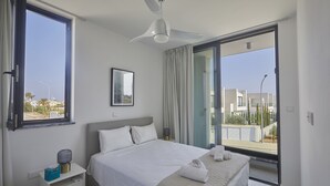 Un dormitorio moderno con una cama grande, ventilador de techo, mesita de noche, ventana y vista al exterior.