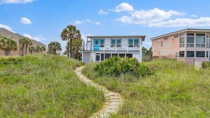 A pathway leading to a modern two-story building with a balcony, surrounded by lush greenery and palm trees. - Courtyard Home with Fire Pit & Fenced Backyard (St. Augustine)