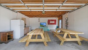 A covered outdoor area with a wooden table and benches, a refrigerator, and a television.