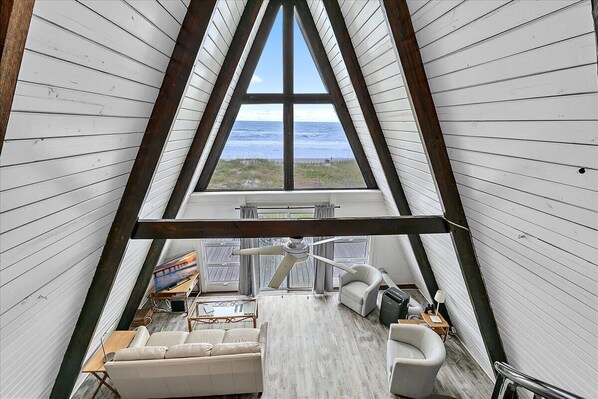 Un habitación estilo loft con techo alto, vigas de madera, un ventanal con vista al océano, un ventilador de techo y un sofá.