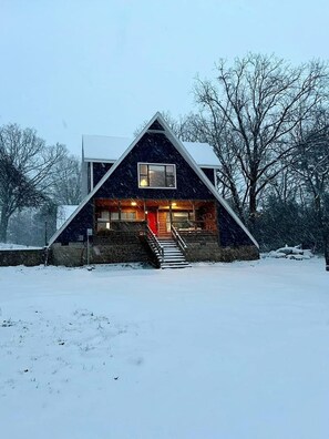A modern, triangular-shaped cabin with a stone foundation, a wooden front porch, and a staircase leading to the entrance.
