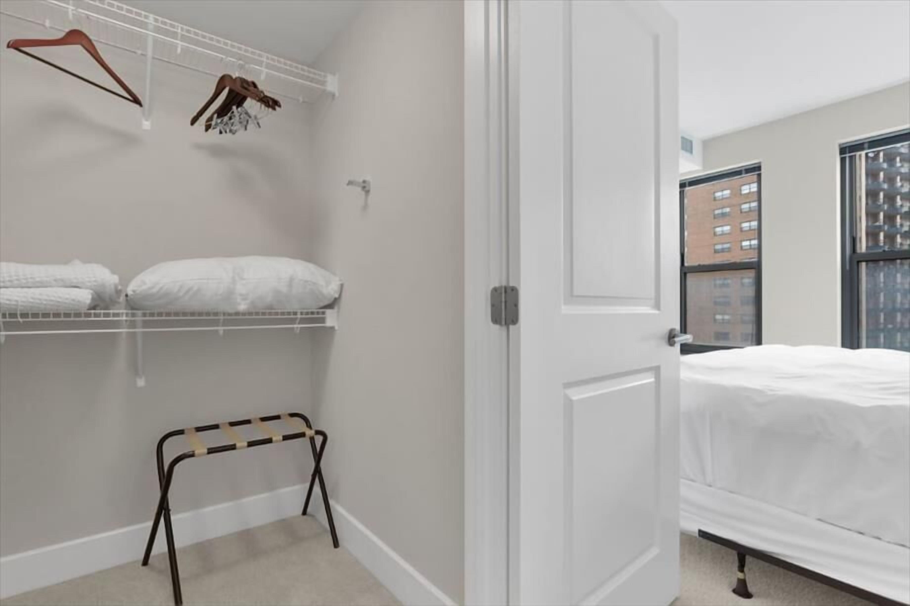 Une chambre à coucher avec un lit, une porte blanche, une armoire où sont suspendus des vêtements et un petit tabouret.