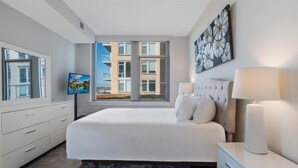 A modern bedroom with a large bed, a TV, a window with a city view, and a painting on the wall. - Mins to Lake Anne Plaza | In/Outdoor Yogs | Reston Town Center (Reston)