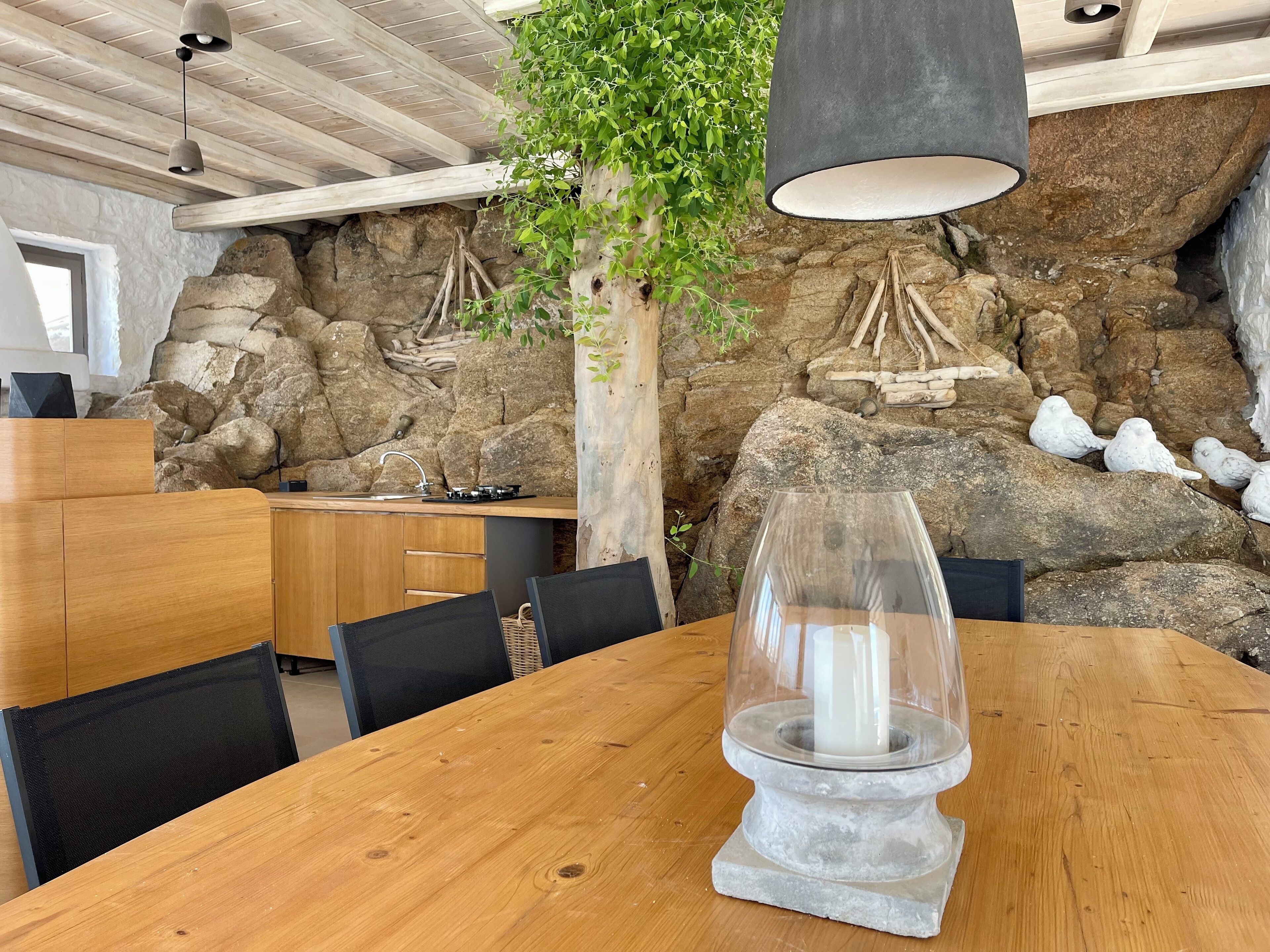 Une table à manger en bois, surmontée d’un porte-bougie en verre transparent placé au centre, est entourée de chaises noires. Un mur de pierre, avec une formation rocheuse et une plante suspendue, se trouve à l’arrière-plan.